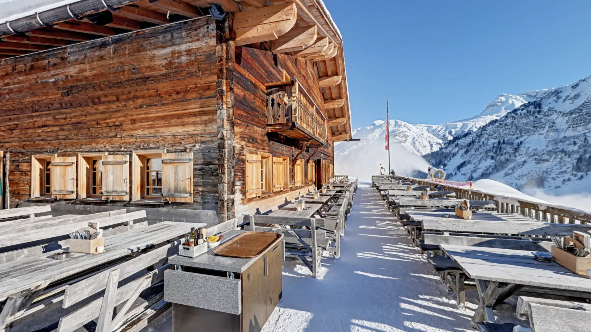 Holzhütte mit Tischen vor schneebedeckten Bergen.