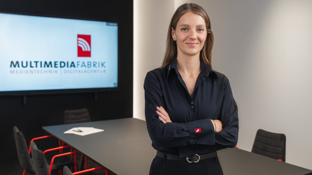 Frau in Büro mit Bildschirm im Hintergrund.