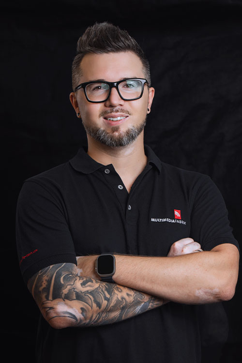 Mann mit Brille, Tattoos und schwarzem Shirt lächelt.