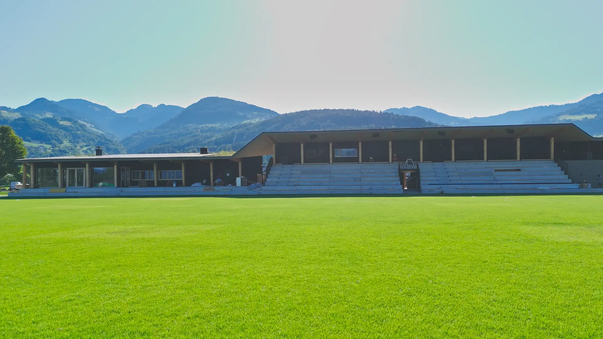Fußballplatz mit Tribüne und Bergen im Hintergrund.