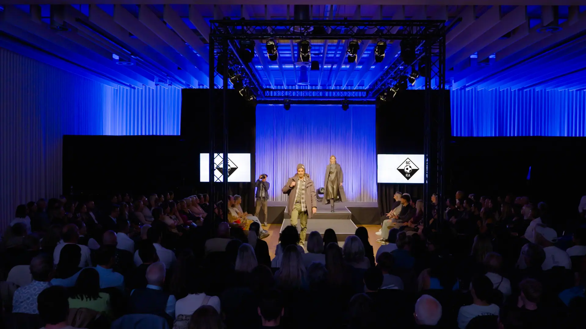 Modenschau auf beleuchtetem Laufsteg mit Publikum.