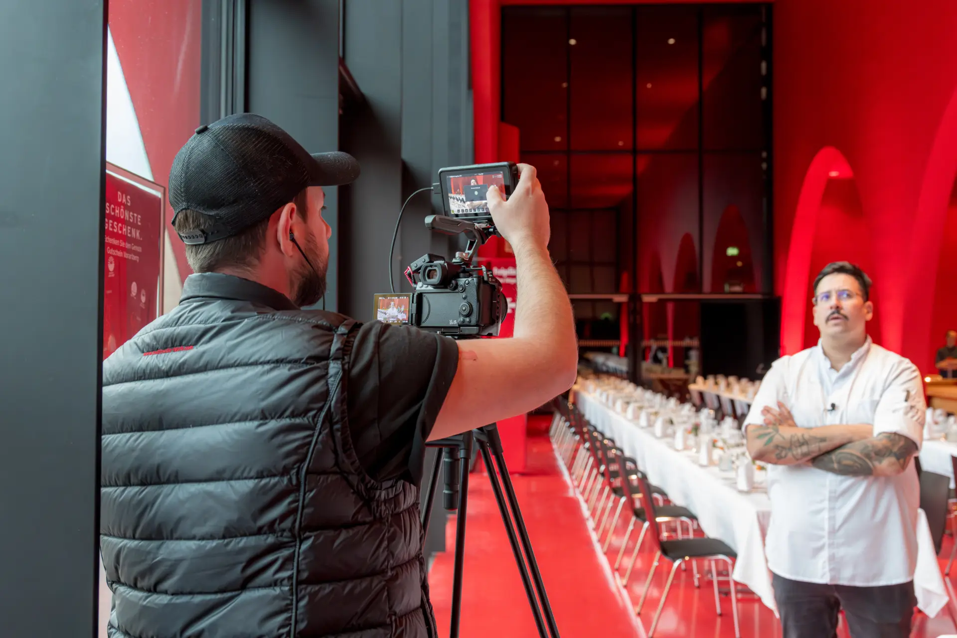Kameramann filmt Person in rotem Raum mit langer Tafel.