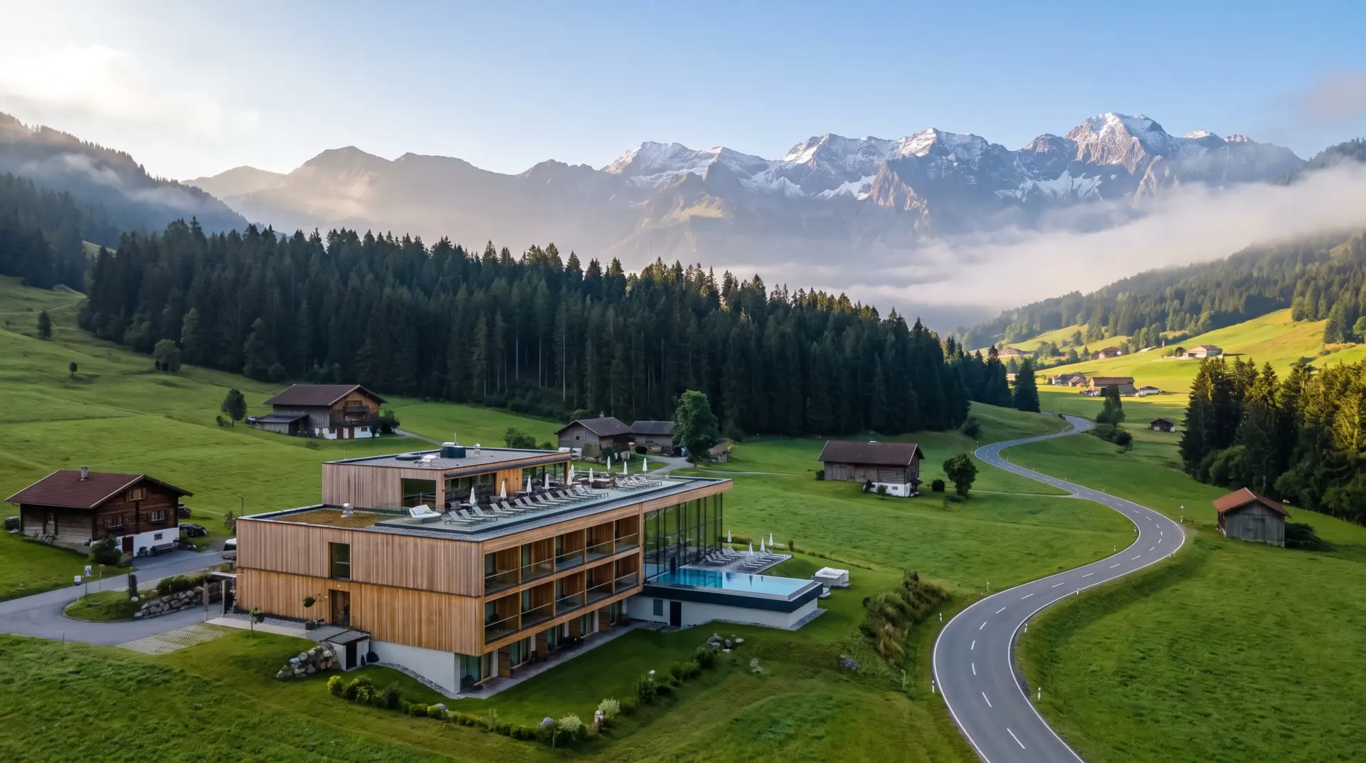 Drohnenaufnahme eines modernen Hotelresorts in einem grünen Alpental mit Dachterrasse, Wiesen und Bergrücken im Hintergrund.