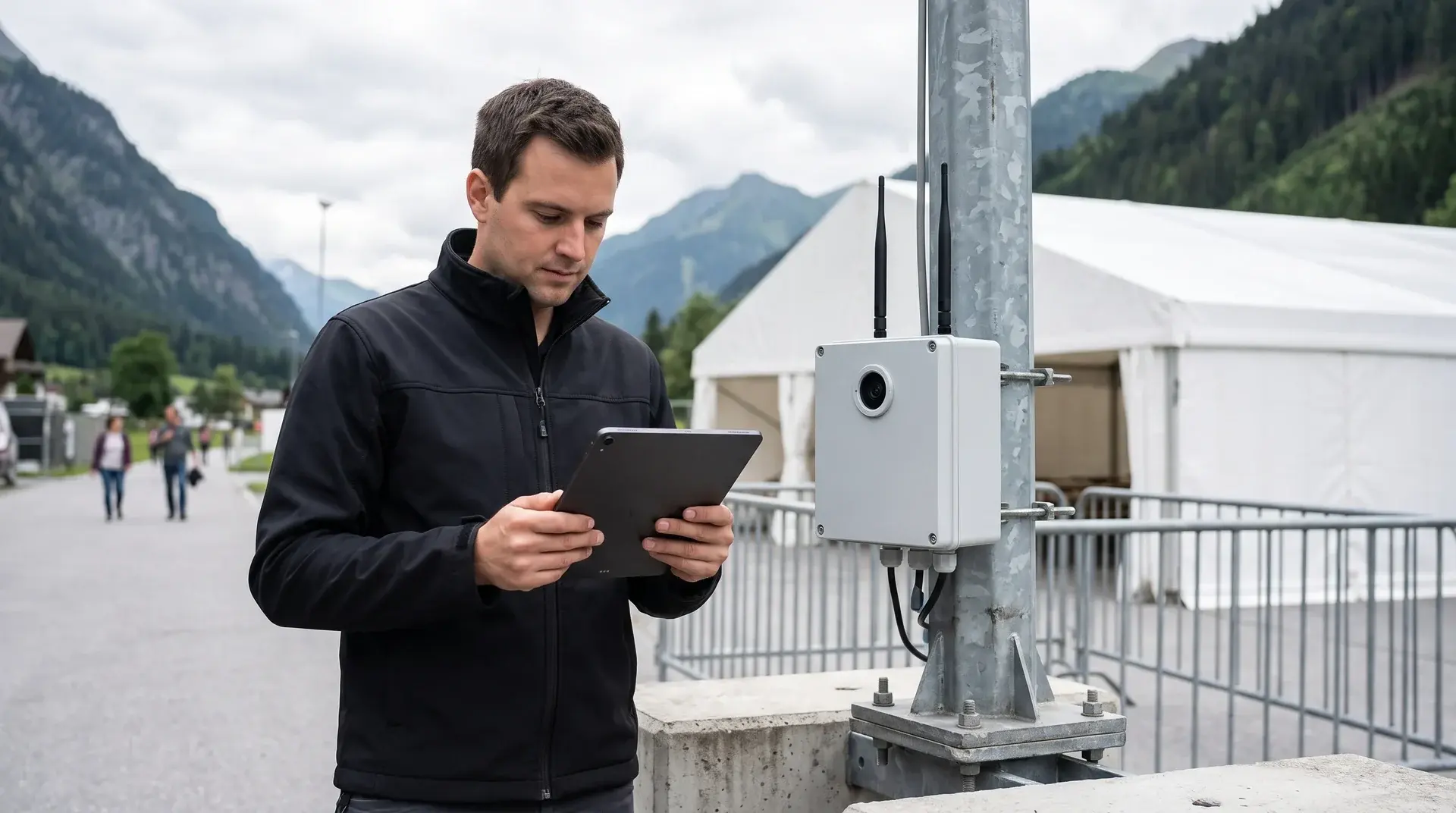 Techniker mit Tablet neben einer Kamera mit grauem Gehäuse und zwei Antennen an einem Mast, dahinter ein Festzelt